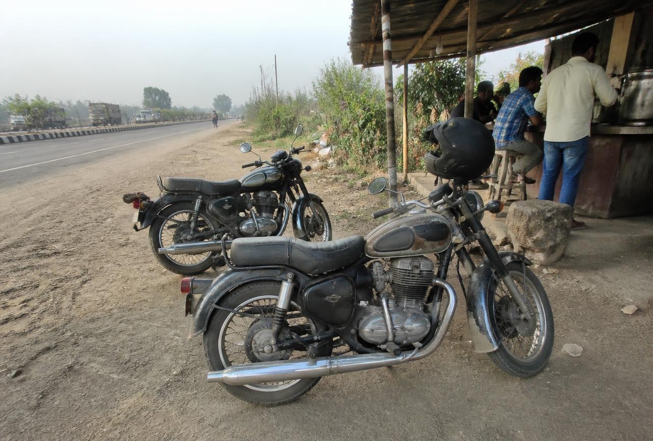 Two retro classic motorcycles parked on gravel outside a roadside tea stall on an Indian highway