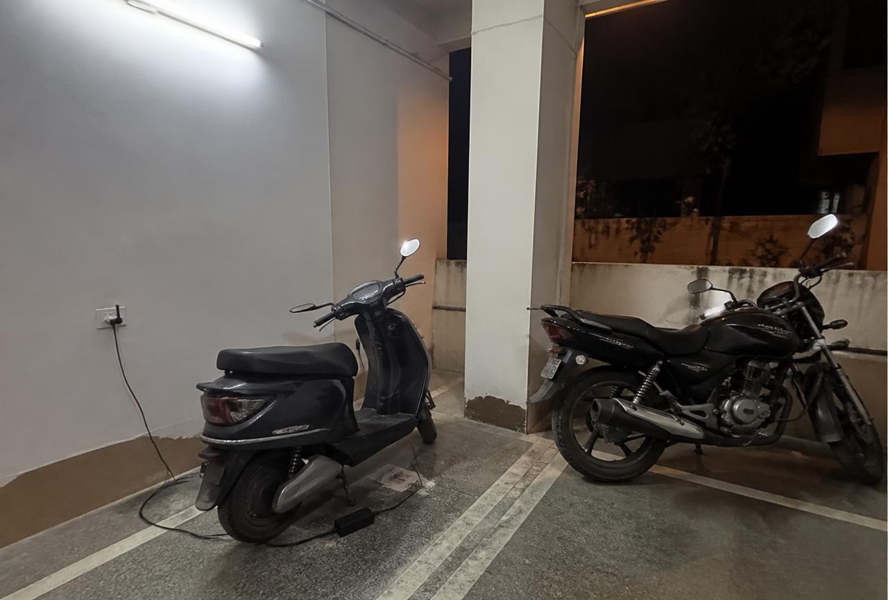 Electric scooter charging from a wall socket next to a petrol commuter motorcycle in an Indian apartment parking area at night